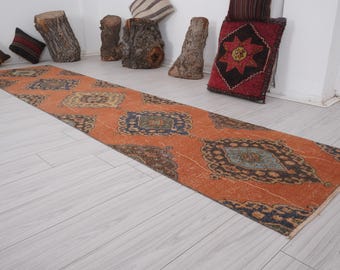 May include: A long, rectangular, orange runner rug with a geometric pattern in shades of blue, brown, and cream. The rug has a fringed edge and is laid out on a light wood floor.