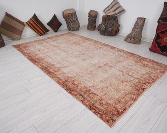 Op de afbeelding: Een groot, rechthoekig, versleten tapijt met een vervaagde perzik- en roestkleurenpalet. Het tapijt heeft een gedetailleerde rand en een versleten, vintage esthetiek. Decoratieve kussens en houten accenten omringen het tapijt.