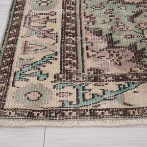 May include: Close-up of a vintage rug with a distressed design. The rug features a beige background with faded patterns in shades of brown, teal, and lavender. The border is a darker brown with a fringed edge, showcasing a classic, antique aesthetic.