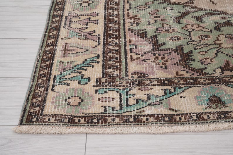 May include: Close-up of a vintage rug with a distressed design. The rug features a beige background with faded patterns in shades of brown, teal, and lavender. The border is a darker brown with a fringed edge, showcasing a classic, antique aesthetic.