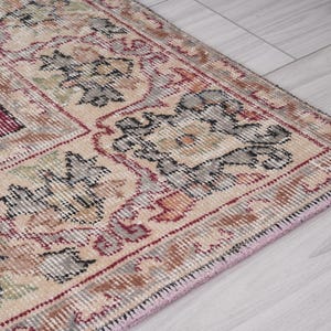 May include: Close-up of a beige rug with intricate floral patterns in shades of red, black, and gray. The rug has a pink fringe border.