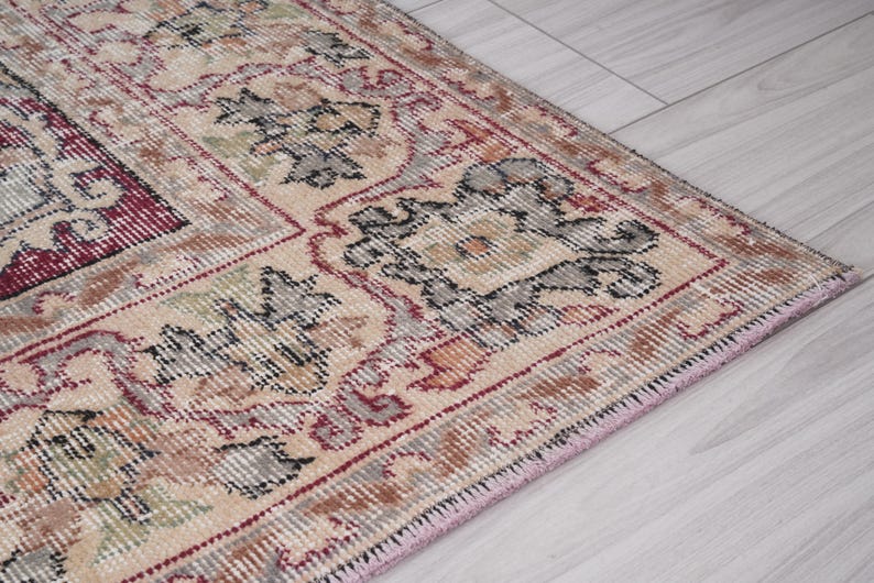 May include: Close-up of a beige rug with intricate floral patterns in shades of red, black, and gray. The rug has a pink fringe border.