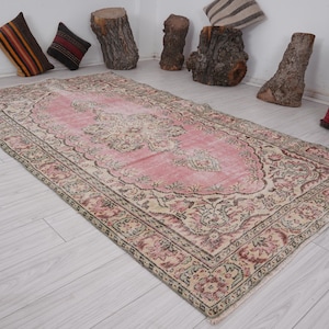 May include: A rectangular, vintage-style rug with a pink central field and ornate, floral-inspired patterns in cream, brown, and burgundy. The rug has a distressed appearance, suggesting age and use. The border features a detailed, repeating design.
