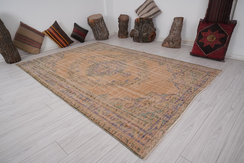 Pu&ograve; includere: Un grande tappeto rettangolare dall'aspetto invecchiato, con un motivo a medaglione centrale nei toni del beige, blu e verde. Il tappeto ha un aspetto sbiadito, che suggerisce uno stile antico o vintage. Cuscini decorativi e elementi in legno sullo sfondo.