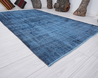 May include: A rectangular, distressed blue rug with a faded appearance. The rug has a dark blue border and a lighter blue center, creating a textured look. The rug is placed on a light-colored wooden floor.