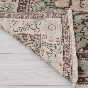 May include: A distressed vintage rug with a beige base and detailed patterns in brown, green, and teal. The rug's edges are fringed, enhancing its aged appearance. The design suggests a traditional style.