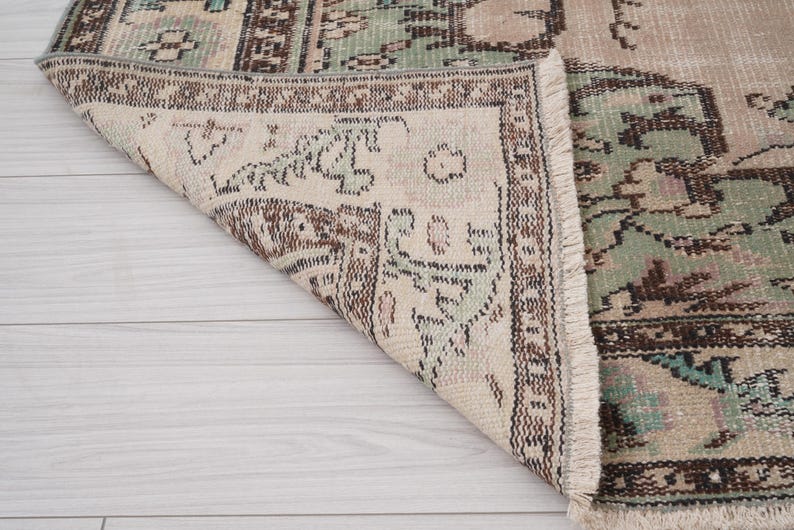 May include: A distressed vintage rug with a beige base and detailed patterns in brown, green, and teal. The rug's edges are fringed, enhancing its aged appearance. The design suggests a traditional style.