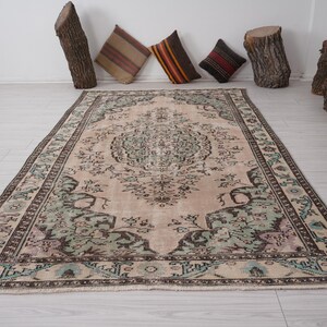 May include: A rectangular rug in beige with an elaborate floral design. The rug has a central medallion and a border with green, blue, and brown details. Decorative cushions and wood stumps are in the background.