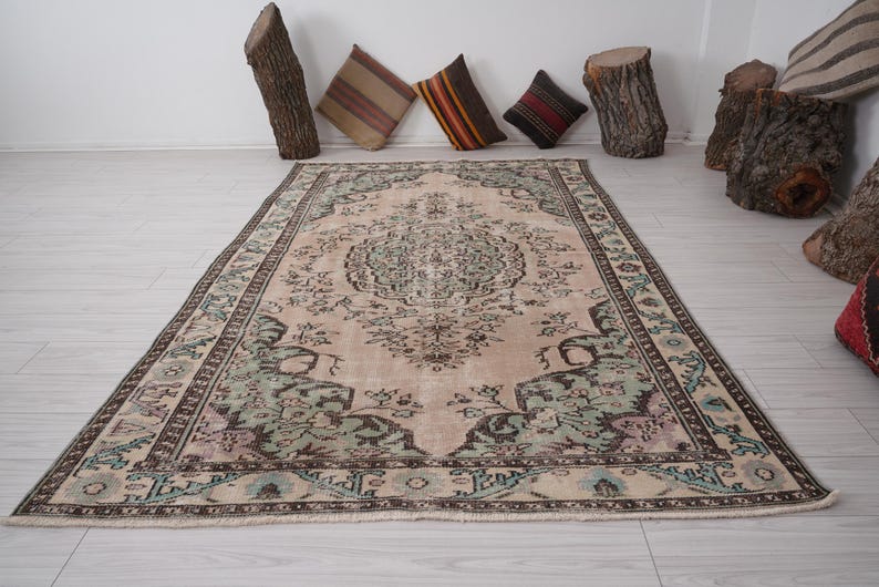 May include: A rectangular rug in beige with an elaborate floral design. The rug has a central medallion and a border with green, blue, and brown details. Decorative cushions and wood stumps are in the background.