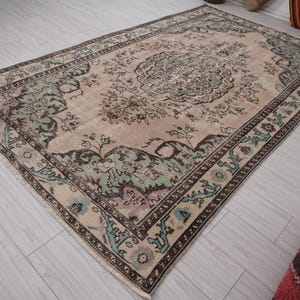 May include: A rectangular rug with a central medallion design in beige, green, and brown. The rug has an ornate floral pattern and a border with geometric and floral elements. It is placed on a light-coloured wooden floor.