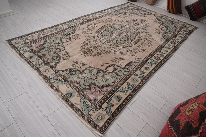 May include: A rectangular rug with a central medallion design in beige, green, and brown. The rug has an ornate floral pattern and a border with geometric and floral elements. It is placed on a light-coloured wooden floor.