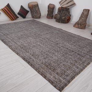 Può includere: Un tappeto rettangolare grigio con una superficie strutturata. Il tappeto ha un bordo scuro ed è posizionato su un pavimento in legno chiaro. Cuscini decorativi e accenti di tronchi di legno sullo sfondo.