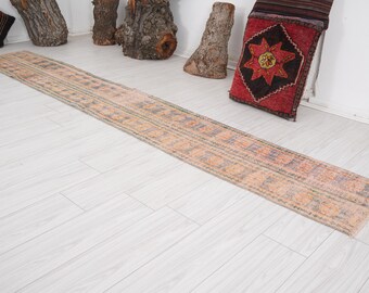May include: A long, rectangular rug with a light pink and cream background and a geometric pattern in shades of blue, green, and orange. The rug is laid out on a light wood floor.