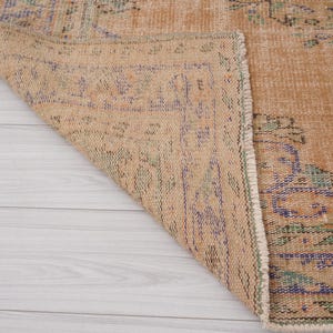 Pu&ograve; includere: Una sezione di un tappeto vintage con una palette di colori beige e sbiaditi. Il tappeto presenta motivi intricati nelle tonalit&agrave; di blu, verde e viola. L'angolo del tappeto &egrave; ripiegato, rivelando il lato inferiore e la frangia dettagliata.