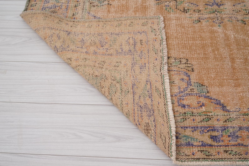 Pu&ograve; includere: Una sezione di un tappeto vintage con una palette di colori beige e sbiaditi. Il tappeto presenta motivi intricati nelle tonalit&agrave; di blu, verde e viola. L'angolo del tappeto &egrave; ripiegato, rivelando il lato inferiore e la frangia dettagliata.