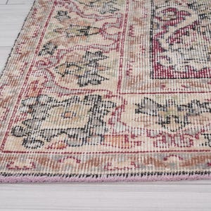 May include: A vintage rug with a cream background and intricate floral patterns in shades of red, black, and gray. The rug has a pink fringe border.