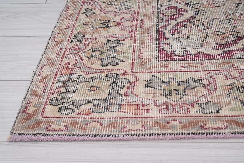 May include: A vintage rug with a cream background and intricate floral patterns in shades of red, black, and gray. The rug has a pink fringe border.