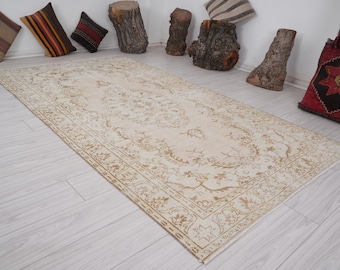Op de afbeelding: Een rechthoekig, beige tapijt met een ingewikkeld, bloemenpatroon. Het tapijt heeft een gedetailleerde rand en ligt op een lichtgekleurde houten vloer. Decoratieve kussens en houten schijven staan op de achtergrond.