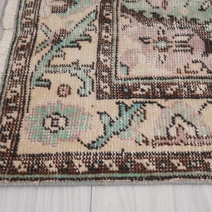 May include: Detail of a vintage rug with a distressed appearance. The rug has a beige base with floral and geometric patterns in brown, teal, and mauve. The rug's edge is fringed.