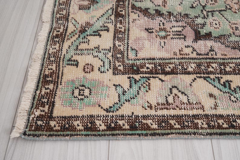 May include: Detail of a vintage rug with a distressed appearance. The rug has a beige base with floral and geometric patterns in brown, teal, and mauve. The rug's edge is fringed.