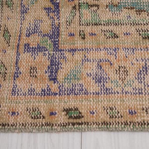 Pu&ograve; includere: Primo piano di una sezione di un tappeto vintage dall'aspetto usurato e sbiadito. Il tappeto presenta uno sfondo beige con motivi intricati in blu, arancione, verde e viola. I bordi hanno un dettaglio a frange.
