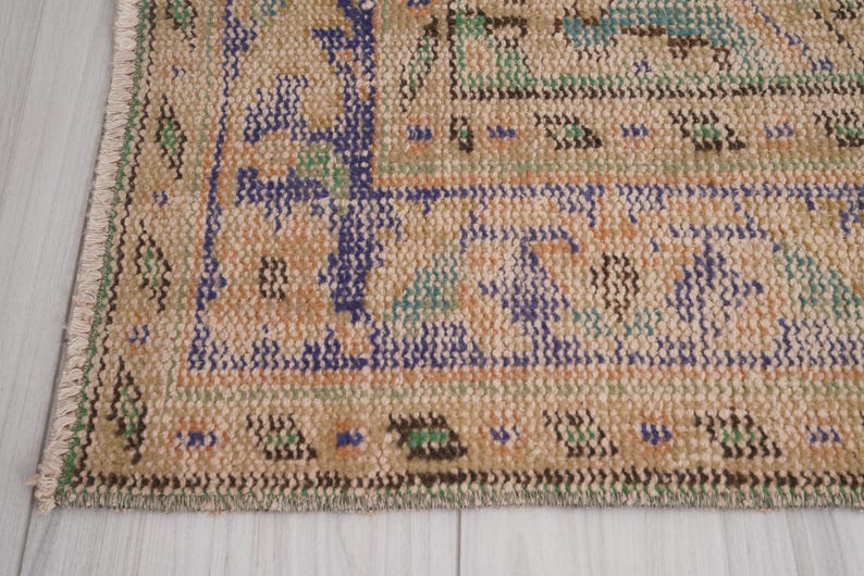Pu&ograve; includere: Primo piano di una sezione di un tappeto vintage dall'aspetto usurato e sbiadito. Il tappeto presenta uno sfondo beige con motivi intricati in blu, arancione, verde e viola. I bordi hanno un dettaglio a frange.