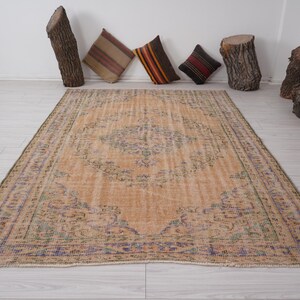 Pu&ograve; includere: Un tappeto rettangolare dall'aspetto invecchiato, con un motivo a medaglione centrale nei toni del beige, blu e verde. Il tappeto &egrave; posato su un pavimento in legno chiaro. Cuscini decorativi e elementi in legno di tronchi sullo sfondo.