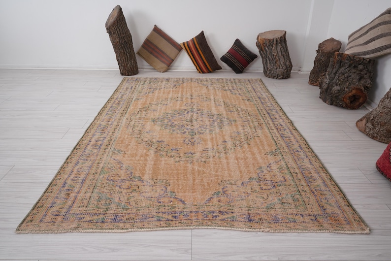 Pu&ograve; includere: Un tappeto rettangolare dall'aspetto invecchiato, con un motivo a medaglione centrale nei toni del beige, blu e verde. Il tappeto &egrave; posato su un pavimento in legno chiaro. Cuscini decorativi e elementi in legno di tronchi sullo sfondo.