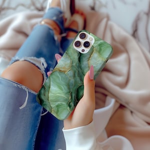 May include: Green and gold marble-patterned phone case. The case has a glossy finish and a gold camera ring. The case is held in a hand with long pink nails. The background shows blue jeans and a soft, neutral-colored blanket.