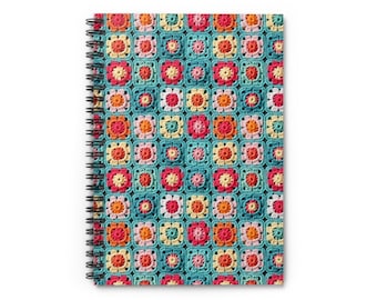 Pode incluir: Um caderno de espiral com um padrão de crochê colorido na capa. O padrão apresenta quadrados de diferentes cores, incluindo rosa, laranja, amarelo e azul, sobre um fundo turquesa.