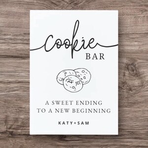 Peut inclure: Panneau blanc avec l'inscription "Cookie Bar" en noir, orné d'un dessin de trois biscuits. Le texte en dessous indique "A Sweet Ending to a New Beginning" et les noms "Katy + Sam".