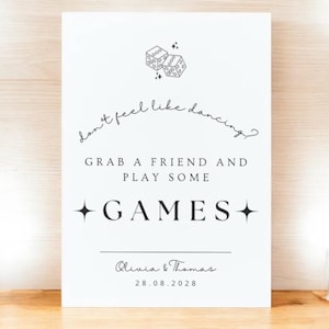 Op de afbeelding: Een wit bord met zwarte tekst en dobbelsteenillustraties. De tekst luidt: "Geen zin om te dansen? Pak een vriend en speel wat spelletjes." Daaronder staat "Olivia & Thomas" en de datum "28.08.2028".