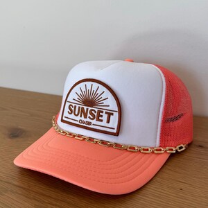 May include: A trucker hat with a white front panel and a coral-colored brim and mesh back. The hat features a brown patch with the words "SUNSET CHASER" in a stylized font, and a gold chain detail.