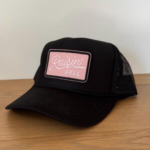 May include: Black trucker hat with a pink rectangular patch. The patch features the words "Raisin' Hell" in a cursive font. The hat has a mesh back and a curved brim.