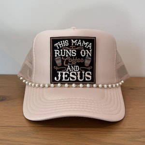 May include: A light pink trucker hat with a pearl beaded brim. The hat features a black square patch with the text "THIS MAMA RUNS ON Coffee AND JESUS" in white and brown. The hat has a mesh back.