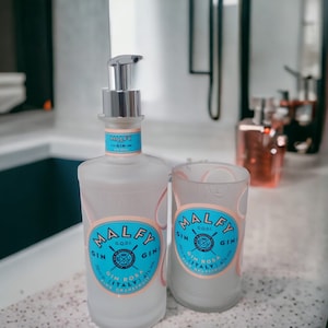 Könnte beinhalten: Zwei milchglasartige Flaschen Malfy Gin, eine mit einer Pumpspender. Beide Flaschen haben ein blaues und weißes Etikett mit dem Text "Malfy Gin G.Q.D.I. Gin Rosa (Italien) 41% vol. Rosa Grapefruit".