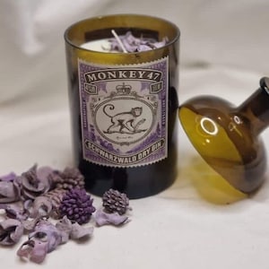 Può includere: Un barattolo di vetro marrone con una candela di cera bianca al suo interno. Il barattolo ha un'etichetta che dice "MONKEY 47 SCHWARZWALD DRY GIN". Il barattolo è appoggiato su una superficie bianca accanto a un tappo di bottiglia di vetro marrone con un tappo di sughero.