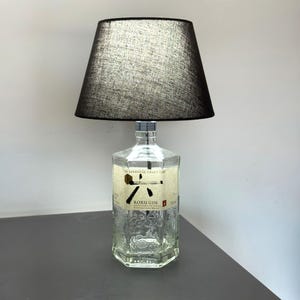 Könnte beinhalten: Eine einzigartige Tischlampe aus einer recycelten Roku Gin-Flasche. Der klare Glasflaschenfuß trägt einen schwarzen Stofflampenschirm. Die Flasche hat ein Etikett mit japanischem Text und einem geometrischen Design.