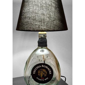 Könnte beinhalten: Eine Tischlampe aus einer durchsichtigen Glasflasche mit einem schwarzen Etikett, auf dem "Vecchia Romagna Etichetta Nera 1820" steht. Die Lampe hat einen schwarzen Stoffschirm.