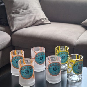 Puede incluir: Seis vasos de chupito transparentes con el logo "MALFY GIN" en verde azulado, naranja y blanco. Los vasos son cilíndricos y esmerilados en la base. El fondo incluye una mesa oscura y un sofá gris.