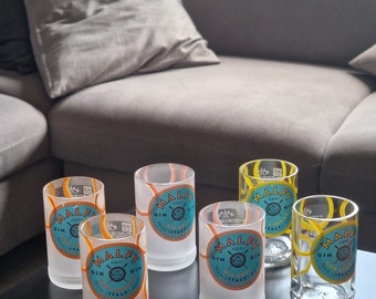 Könnte beinhalten: Sechs klare Schnapsgläser mit dem "MALFY GIN" Logo in Türkis, Orange und Weiß. Die Gläser sind zylindrisch und am Boden mattiert. Der Hintergrund umfasst einen dunklen Tisch und ein graues Sofa.