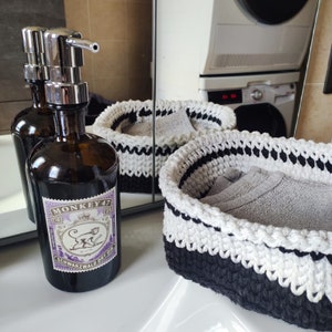 Può includere: Una bottiglia di vetro trasparente con un tappo a pompa nero e argento. La bottiglia è etichettata "Monkey 47 Schwarzwald Dry Gin". La bottiglia è appoggiata su un bancone bianco accanto a due cesti in crochet bianco e nero.