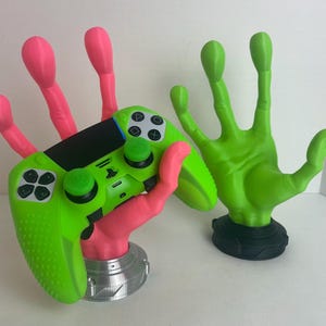 Könnte beinhalten: Eine grün und pink 3D-gedruckte Hand hält einen grünen Videospiel-Controller. Die Hand hält den Controller so, dass es aussieht, als würde sie ein Spiel spielen. Der Controller hat ein schwarz-weißes Design und ist aus Kunststoff. Die Hand befindet sich auf einer weißen Oberfläche.