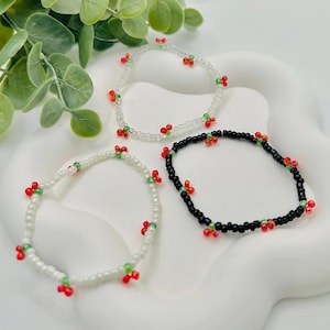 Op de afbeelding: Drie kralenarmbanden met rode en groene kersenontwerpen. Een armband is wit, een is zwart en een is helder.