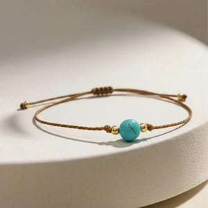 Könnte beinhalten: Ein minimalistisches Armband mit einer leuchtenden türkisfarbenen Perle als Mittelpunkt, ergänzt durch kleine goldfarbene Perlen. Das Armband ist an einem braunen Kordelband befestigt, mit verstellbaren Knoten. Ein schlichtes und elegantes Schmuckstück.