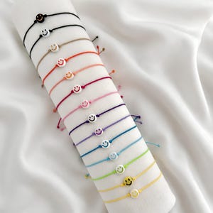 Pode incluir: Uma coleção de treze pulseiras de cordão ajustáveis, cada uma com uma conta branca com um rosto sorridente colorido. As pulseiras são exibidas em uma fileira, mostrando uma variedade de cores de cordão, incluindo preto, bege, vermelho, rosa, roxo, azul, verde e amarelo.