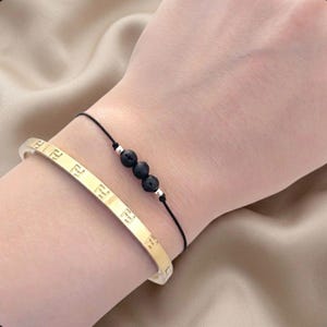 Könnte beinhalten: Zwei Armbänder an einem Handgelenk. Eines ist ein goldfarbenes Armreif mit sich wiederholenden geprägten Buchstaben. Das andere ist ein schwarzes Kordelarmband mit drei schwarzen Perlen und silberfarbenen Akzenten. Die Armbänder werden auf hellem Hautton getragen.