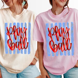 May include: Two short-sleeved t-shirts, one cream and one pink, each featuring the words "Play Ball" in red script over blue vertical stripes. The shirts are casual and appear to be made of a soft material.