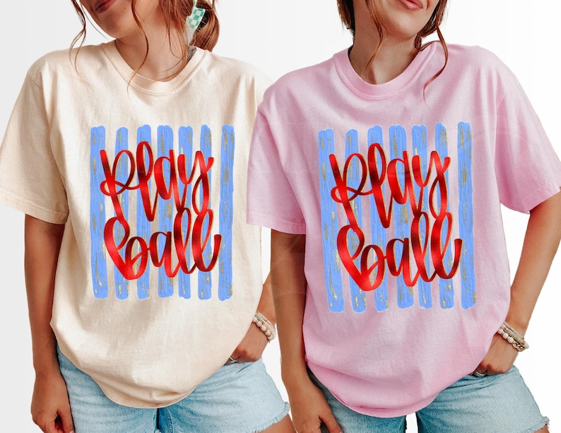 May include: Two short-sleeved t-shirts, one cream and one pink, each featuring the words "Play Ball" in red script over blue vertical stripes. The shirts are casual and appear to be made of a soft material.