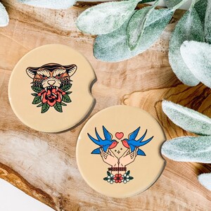 May include: Two round beige coasters with colorful illustrations. One coaster features a tiger with a red rose in its mouth. The other coaster features two blue birds with a red heart between them.